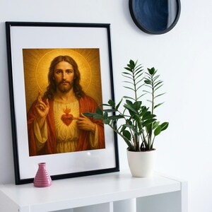 May include: Framed religious artwork featuring a portrait of Jesus Christ. The image shows Jesus with long hair, a beard, and a halo, pointing to his heart. The artwork is framed in black and displayed on a white shelf with a small pink vase and a potted plant.