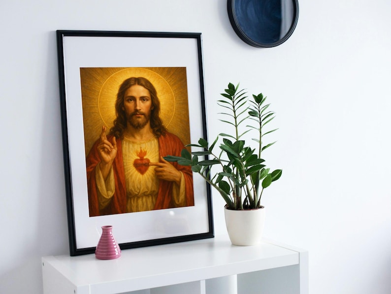 May include: Framed religious artwork featuring a portrait of Jesus Christ. The image shows Jesus with long hair, a beard, and a halo, pointing to his heart. The artwork is framed in black and displayed on a white shelf with a small pink vase and a potted plant.