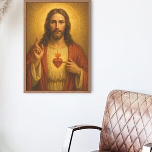 May include: A framed religious portrait depicts a man with long brown hair, a beard, and a halo. He wears a red robe and points to a flaming heart on his chest. The artwork is displayed on a white wall near a brown leather chair.