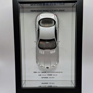 May include: A silver Mercedes Benz SLR McLaren model car displayed in a black shadow box frame. The white background features text: MERCEDES BENZ SLR MCLAREN, YEAR: 2003, ENGINE: 5.4L SUPERCHARGED V8, 0-60: 3.5 sec, POWER: 624 HP, TOP SPEED: 200 MPH.