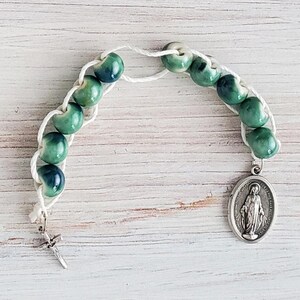 May include: A green and white rosary with a silver charm of the Virgin Mary. The rosary is made of small, round beads and is strung on a white cord.