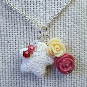 Könnte beinhalten: Eine Halskette mit einem weißen sternförmigen Anhänger, der mit einem weißen, gelben und rosafarbenen Blumenanhänger verziert ist. Der Anhänger ist aus Lavastein gefertigt und hat einen funkelnden roten Kristall als Akzent.