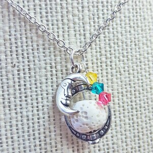 Könnte beinhalten: Eine silberne Halskette mit einem Halbmond-Anhänger, der eine weiße Lavasteinkugel umschließt. Der Anhänger ist mit kleinen bunten Kristallen in Gelb, Grün und Rosa verziert.