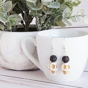 Puede incluir: Un par de pendientes colgantes con diseño de calavera. Cada pendiente tiene una cuenta de calavera blanca con acabado crema, rematada con una cuenta negra texturizada. Los pendientes cuelgan de ganchos plateados.