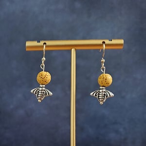 Könnte beinhalten: Ein Paar goldene Bienenohrringe mit gelben Lavasteinen, die an einem goldenen Ständer auf einer Holzoberfläche hängen.