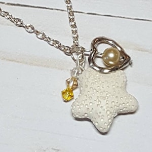 Könnte beinhalten: Eine silberne Kettenkette mit einem weißen Seestern-Anhänger, einer kleinen Perle in einem silbernen Herz-Anhänger und einem gelben Kristall-Anhänger.