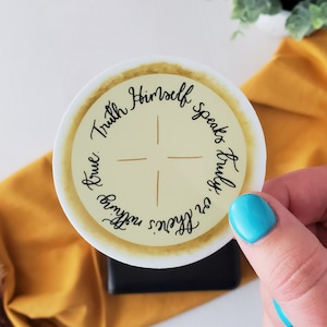 Pode incluir: Um autocolante redondo com o texto "Truth Himself speaks truly, or there's nothing true." O autocolante tem um centro amarelo claro com um desenho de cruz e uma borda creme. O texto está em escrita preta.