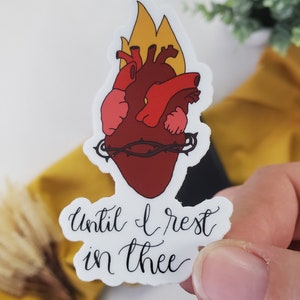 May include: A sticker depicting a stylized anatomical heart in red and pink, encircled by a thorny crown. Yellow and orange flames rise above the heart. The script reads "Until I rest in thee."