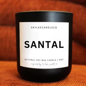 May include: A black candle with a white label featuring the word "SANTAL" in bold letters. The label also includes the text "SKYLASCANDLECO" and "NATURAL SOY WAX CANDLE | 8OZ". The candle is set against a warm, orange-brown background.