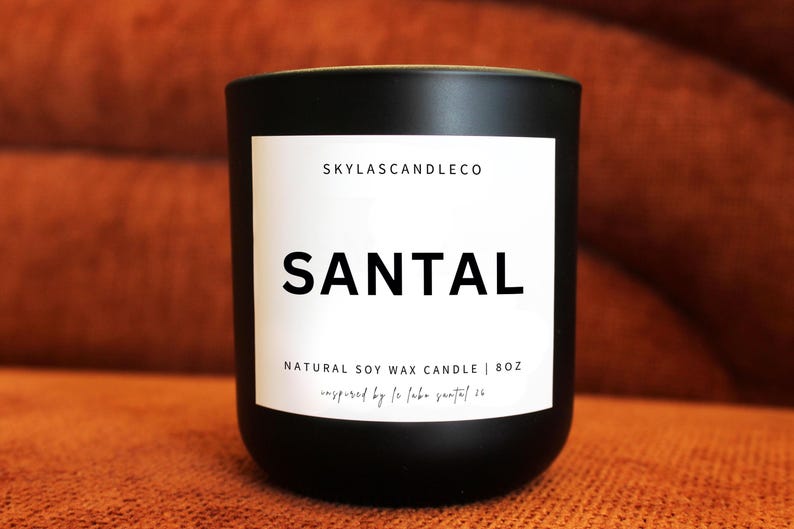 May include: A black candle with a white label featuring the word "SANTAL" in bold letters. The label also includes the text "SKYLASCANDLECO" and "NATURAL SOY WAX CANDLE | 8OZ". The candle is set against a warm, orange-brown background.
