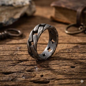 May include: A silver-toned ring with a chain link design. The ring has a textured, oxidized finish and features diamond-shaped cutouts. The ring is displayed on a rustic wooden surface, with other vintage items in the background.
