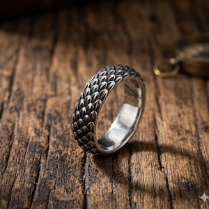 Peut inclure: Une bague en argent avec un motif texturé en forme d'écailles. La bague est en métal brillant et est posée sur un fond en bois rustique. Le design de la bague est détaillé et accrocheur.