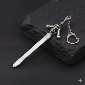 May include: A silver-colored sword-shaped keychain. The keychain features a detailed design of a sword with a skull and crossbones on the hilt. The sword blade is long and pointed, and the keychain includes a ring for attaching keys.