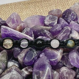 Pode incluir: Uma pulseira de macramé preta com cinco contas de pedras preciosas: transparentes, pretas, rosas e verdes. A pulseira é exibida sobre uma cama de pedras de ametista roxas, com um fundo de serapilheira.