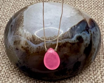 May include: A pink teardrop-shaped pendant necklace on a brown string. The necklace is resting on a brown and white agate stone.