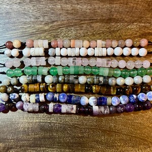 May include: A selection of beaded bracelets in shades of pink, green, yellow, and purple. Each bracelet showcases a different gemstone or bead type, threaded on a black cord. The beads are round, cylindrical, and rectangular in shape.