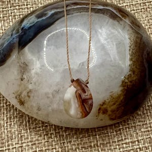 May include: A teardrop-shaped agate pendant with brown and white swirling patterns hangs from a simple brown cord necklace.