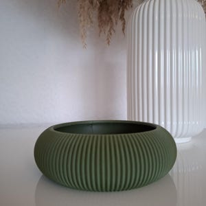 Könnte beinhalten: Ein grüner Keramik-Pflanztopf mit geripptem Design. Der Topf ist rund und hat eine breite Öffnung.