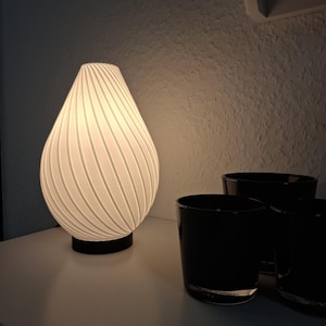 Könnte beinhalten: Eine weiße, gerippte, tropfenförmige Tischlampe mit schwarzem Fuß. Die Lampe ist eingeschaltet und beleuchtet den Raum. Drei schwarze Glaskerzenhalter stehen auf dem Tisch neben der Lampe.