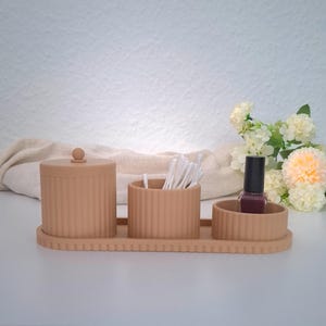 Könnte beinhalten: Ein beiges Badezimmer-Organizer-Set. Das Set enthält ein Gefäß mit Deckel, einen Behälter mit Wattestäbchen und eine kleine Schale mit Nagellack. Alle Teile haben ein geripptes Design und stehen auf einem passenden Tablett. Im Hintergrund sind Blumen.