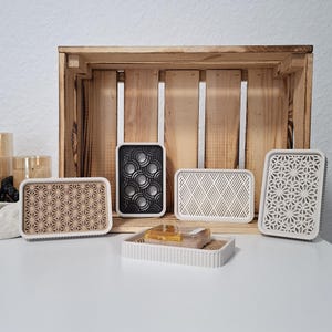 Könnte beinhalten: Eine Sammlung rechteckiger Seifenschalen in verschiedenen Farben und Mustern. Die Schalen werden auf einer weißen Oberfläche vor einer Holzkiste präsentiert. Designs umfassen geometrische und florale Muster, mit einer Seifenschale mit zwei Seifenstücken.