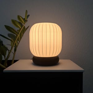 Könnte beinhalten: Eine weiße Tischlampe mit einem gerippten Glasschirm und einem dunklen Holzsockel. Die Lampe ist eingeschaltet und strahlt ein warmes, weißes Licht aus.
