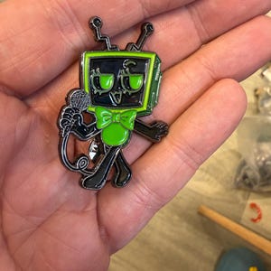 May include: A metal enamel pin featuring a black and lime green television-headed character with antennae, wearing a lime green bow tie, and holding a microphone. The character has large eyes, a wide smile, and thin black limbs with a glossy finish.