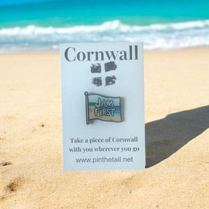 Può includere: Un biglietto bianco con il testo "Cornwall" in lettere nere. Il biglietto presenta anche una piccola spilla smaltata colorata che rappresenta una bandiera con il testo "JAM FIRST" su di essa. Il biglietto è appoggiato su una spiaggia di sabbia con acqua di mare blu sullo sfondo.
