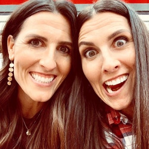 May include: Two women with long brown hair are smiling at the camera. The woman on the left is wearing a gold necklace and earrings. The woman on the right is wearing a red and white plaid shirt.