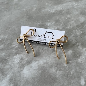 May include: A pair of gold wire earrings shaped like bows. The earrings are on a gray textured surface with a white card that says "Lusted Pearl" and "crafted from vintage materials".
