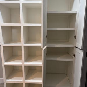 May include: A white wooden storage cabinet with multiple open compartments and shelves. The cabinet has a grid-like design on one side and a door on the other, revealing shelves. The interior is empty, showcasing the clean, white finish.