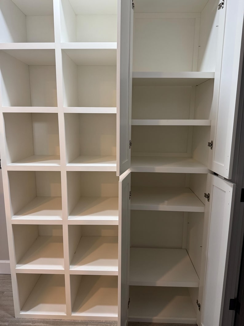 May include: A white wooden storage cabinet with multiple open compartments and shelves. The cabinet has a grid-like design on one side and a door on the other, revealing shelves. The interior is empty, showcasing the clean, white finish.