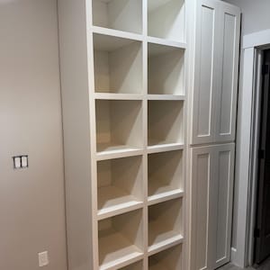 May include: A white built-in storage unit with open cubby shelves and a cabinet door section. The unit is against a light grey wall and sits on a grey tiled floor. The shelves are empty.