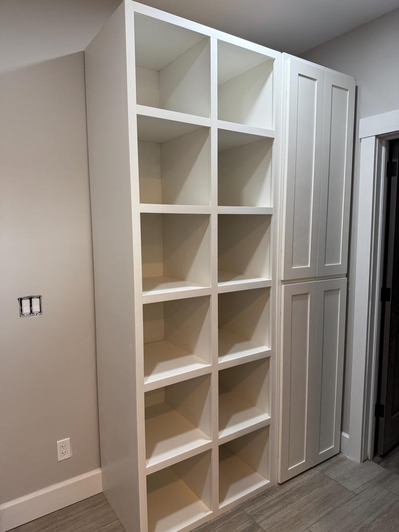 May include: A white built-in storage unit with open cubby shelves and a cabinet door section. The unit is against a light grey wall and sits on a grey tiled floor. The shelves are empty.