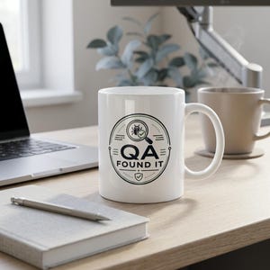 May include: A white ceramic mug featuring a black circular graphic with a magnifying glass over a bug icon, the text "QA FOUND IT", and a small shield icon at the bottom. The mug has a standard handle and a smooth, glossy finish.