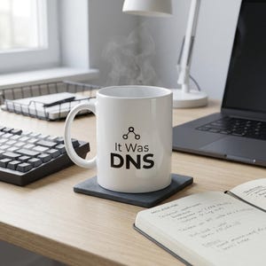Könnte beinhalten: Ein weißer Keramikbecher mit Henkel, der eine schwarze Grafik eines Netzwerkknoten-Symbols über dem Text "It Was DNS" in schwarzer Schrift zeigt. Der Becher ist mit einem Heißgetränk gefüllt, das sichtbaren Dampf aufsteigen lässt.
