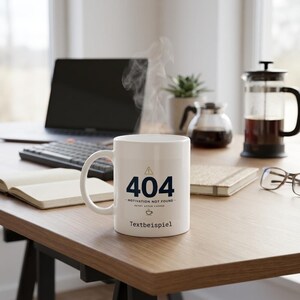 Może przedstawiać: Biały ceramiczny kubek z ciemnoniebieską grafiką błędu 404, napisem Motivation Not Found, Retry After Coffee oraz słowem Textbeispiel wydrukowanym poniżej. Z kubka, który posiada zakrzywione ucho z boku, unosi się para wodna.