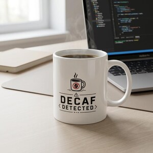 Könnte beinhalten: Ein weißer Keramikbecher gefüllt mit dunklem Kaffee, verziert mit einer schwarzen Grafik einer Kaffeetasse mit einem roten Verbotszeichen über einer Kaffeebohne. Darunter steht der Text DECAF DETECTED in fetten schwarzen Großbuchstaben.