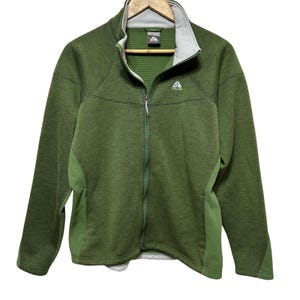 May include: A long-sleeve olive green zip-up jacket featuring a light gray inner collar, a textured grid pattern on the interior, and a small white Nike ACG logo on the upper left chest. The garment includes side pockets and a full-length front zipper.