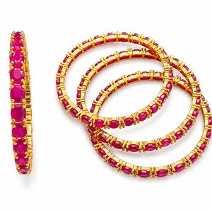 May include: A set of four gold bangles, each featuring a continuous row of vibrant, oval-cut fuchsia-pink stones set in a pronged metal frame. One bangle is displayed vertically on the left, while three are interlocked in a cluster on the right.