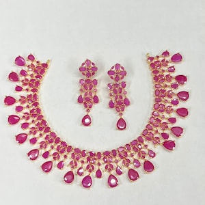 Op de afbeelding: Een goudkleurige ketting en bijpassende oorbellen set. De sieraden hebben talrijke fuchsia-kleurige, druppelvormige edelstenen. De ketting en oorbellen zijn op een witte achtergrond geplaatst, wat het ingewikkelde ontwerp en de levendige kleur laat zien.