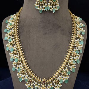 May include: A gold-toned necklace and matching earrings set. The necklace features a central pendant and cascading green teardrop-shaped stones, white pearls, and small red and green accents. The earrings mirror the design.