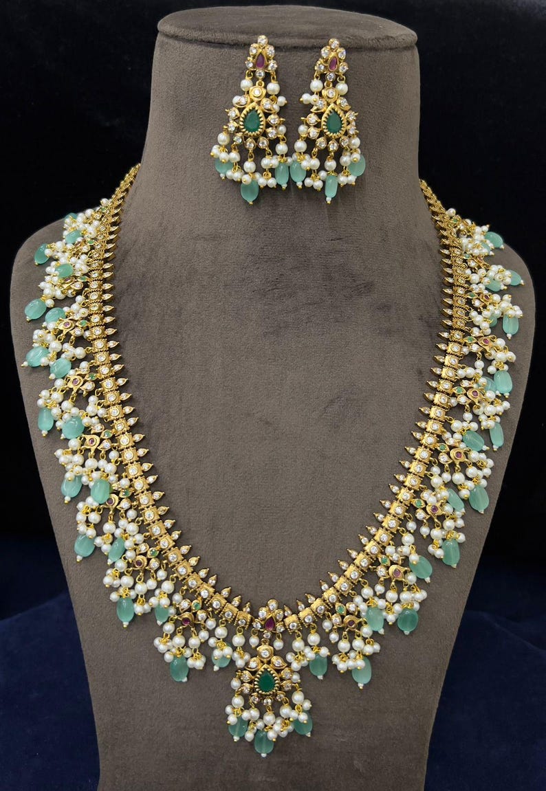 May include: A gold-toned necklace and matching earrings set. The necklace features a central pendant and cascading green teardrop-shaped stones, white pearls, and small red and green accents. The earrings mirror the design.