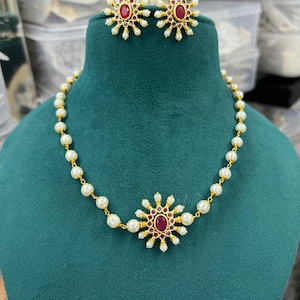 May include: A gold-toned necklace and matching earrings set. The necklace features a central floral design with a ruby-colored stone, surrounded by small pearls. The earrings mirror the design. The set is displayed on a green velvet bust.