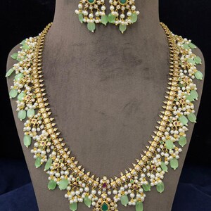 May include: A gold-toned necklace and earring set. The necklace features light green teardrop-shaped stones, small white pearls, and small red and green gemstones. The earrings mirror the necklace design. The jewelry is displayed on a mannequin.
