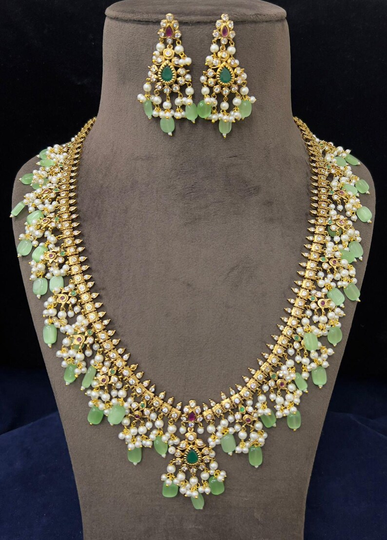 May include: A gold-toned necklace and earring set. The necklace features light green teardrop-shaped stones, small white pearls, and small red and green gemstones. The earrings mirror the necklace design. The jewelry is displayed on a mannequin.