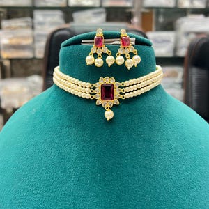 May include: A gold-tone choker necklace and matching earring set featuring three rows of small white pearls. The center of the choker and each earring display a rectangular deep red stone surrounded by clear crystals and small dangling pearls.