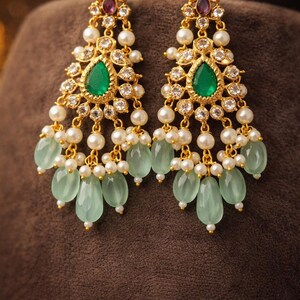May include: Gold-toned chandelier earrings with a teardrop-shaped emerald green stone at the center. The earrings feature white pearls, clear crystals, and light green teardrop-shaped beads. A small purple stone is at the top of each earring.
