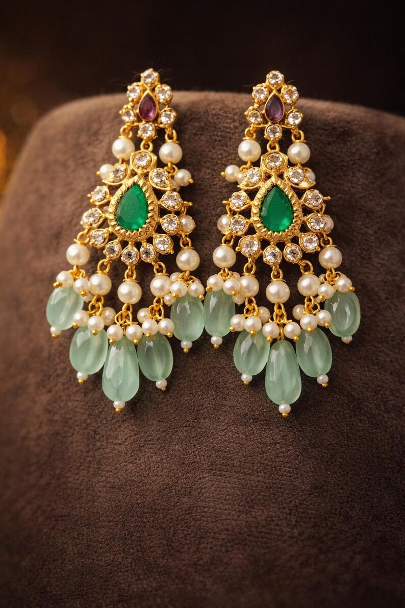 May include: Gold-toned chandelier earrings with a teardrop-shaped emerald green stone at the center. The earrings feature white pearls, clear crystals, and light green teardrop-shaped beads. A small purple stone is at the top of each earring.