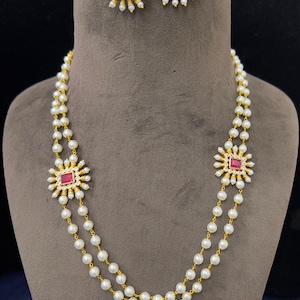 May include: A two-strand pearl necklace and matching earrings set. The necklace features a gold chain with white pearls and a central square red gemstone surrounded by a sunburst design. The earrings have a similar design.